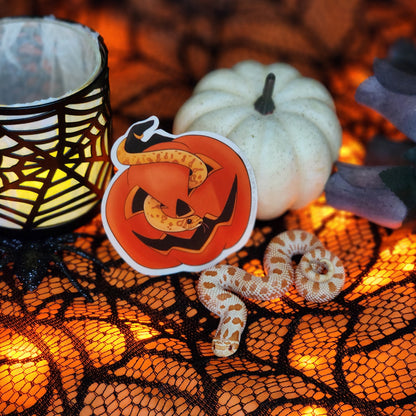 Halloween-themed decor with a lit candle, pumpkin-shaped sticker, and snake on a spiderweb-patterned cloth.